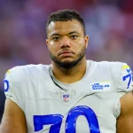 Ein Footballspieler namens Joseph Noteboom trägt ein weißes Trikot der Los Angeles Rams mit der blauen Nummer 70 und gelben Akzenten. Er hat kurze Haare, einen gestutzten Bart und einen ernsten Gesichtsausdruck. Der unscharfe Stadionhintergrund ist in Rot- und Lilatönen gehalten. Diese Beschreibung wurde mit der FootballR KI automatisch generiert.