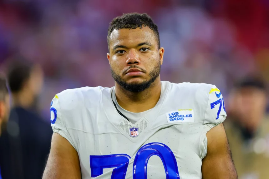 Ein Footballspieler namens Joseph Noteboom trägt ein weißes Trikot der Los Angeles Rams mit der blauen Nummer 70 und gelben Akzenten. Er hat kurze Haare, einen gestutzten Bart und einen ernsten Gesichtsausdruck. Der unscharfe Stadionhintergrund ist in Rot- und Lilatönen gehalten. Diese Beschreibung wurde mit der FootballR KI automatisch generiert.