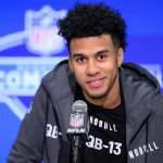 Jordan Travis mit kurzen Locken und Bart lächelt, während er bei einer Pressekonferenz in ein NFL-Mikrofon spricht. Jordan Travis trägt eine dunkle Jacke mit Reißverschluss über einem schwarzen Shirt mit der Aufschrift „QB-13“. Hinter ihm ist ein verschwommenes NFL Combine-Logo zu sehen. Diese Beschreibung wurde mit der FootballR KI automatisch generiert.