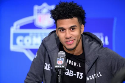 Jordan Travis mit kurzen Locken und Bart lächelt, während er bei einer Pressekonferenz in ein NFL-Mikrofon spricht. Jordan Travis trägt eine dunkle Jacke mit Reißverschluss über einem schwarzen Shirt mit der Aufschrift „QB-13“. Hinter ihm ist ein verschwommenes NFL Combine-Logo zu sehen. Diese Beschreibung wurde mit der FootballR KI automatisch generiert.
