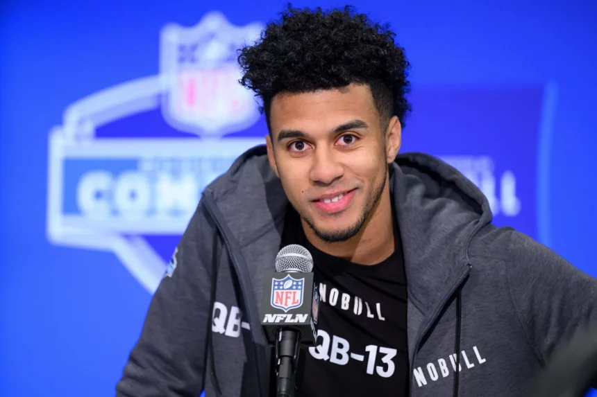 Jordan Travis mit kurzen Locken und Bart lächelt, während er bei einer Pressekonferenz in ein NFL-Mikrofon spricht. Jordan Travis trägt eine dunkle Jacke mit Reißverschluss über einem schwarzen Shirt mit der Aufschrift „QB-13“. Hinter ihm ist ein verschwommenes NFL Combine-Logo zu sehen. Diese Beschreibung wurde mit der FootballR KI automatisch generiert.