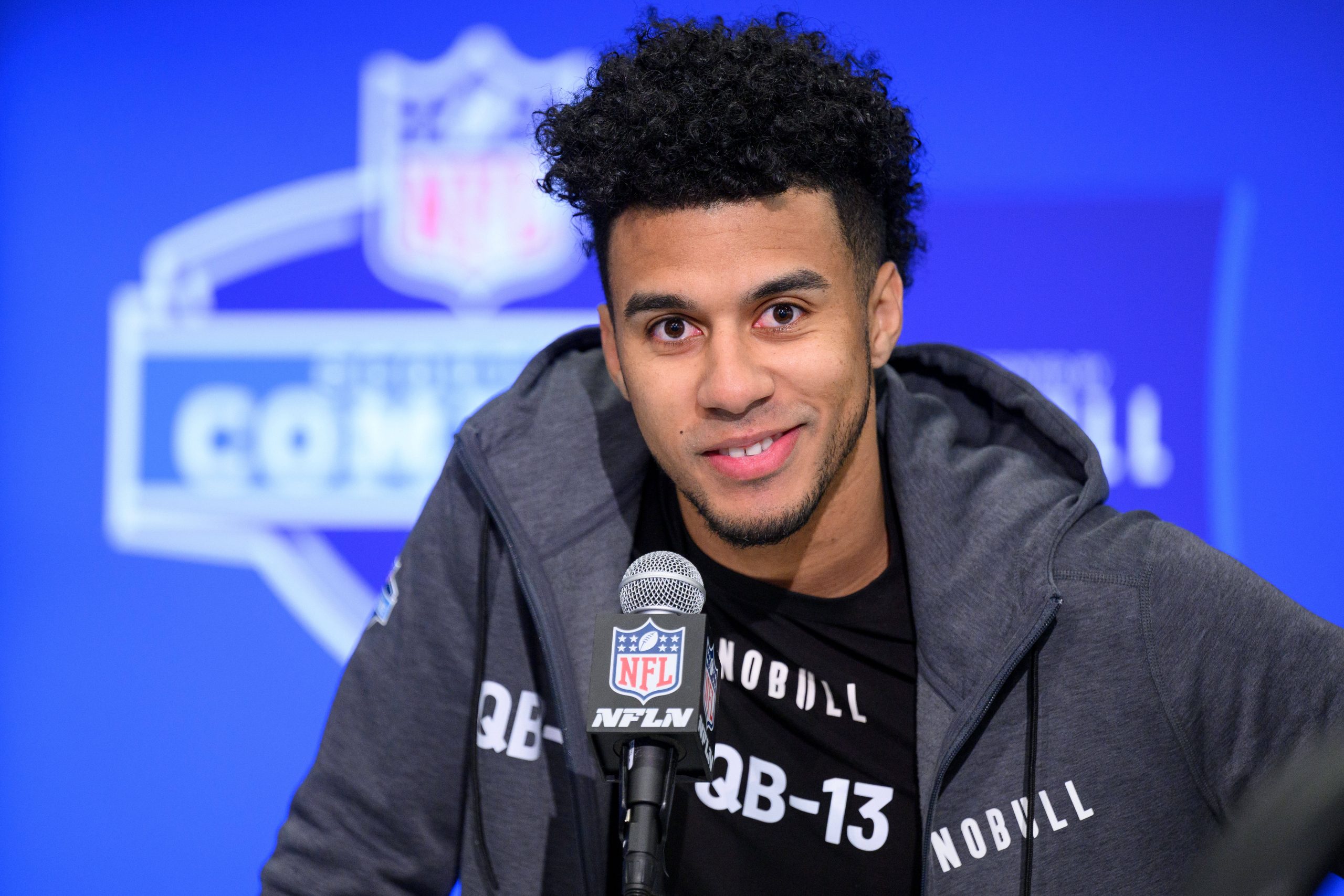 Jordan Travis mit kurzen Locken und Bart lächelt, während er bei einer Pressekonferenz in ein NFL-Mikrofon spricht. Jordan Travis trägt eine dunkle Jacke mit Reißverschluss über einem schwarzen Shirt mit der Aufschrift „QB-13“. Hinter ihm ist ein verschwommenes NFL Combine-Logo zu sehen. Diese Beschreibung wurde mit der FootballR KI automatisch generiert.