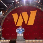 NFL Draft 2027 - Auf einer großen Bühne prangt ein riesiges gelbes „W“-Logo auf rotem Grund. Darunter sind das NFL Draft 2024-Schild und „DETROIT“ zu sehen. Die Bühne ist mit hellen Scheinwerfern beleuchtet. Links neben einem durchsichtigen Podium stehen Menschen; in den Ecken sind Bud-Light-Logos zu sehen. Diese Beschreibung wurde mit der FootballR KI automatisch generiert.
