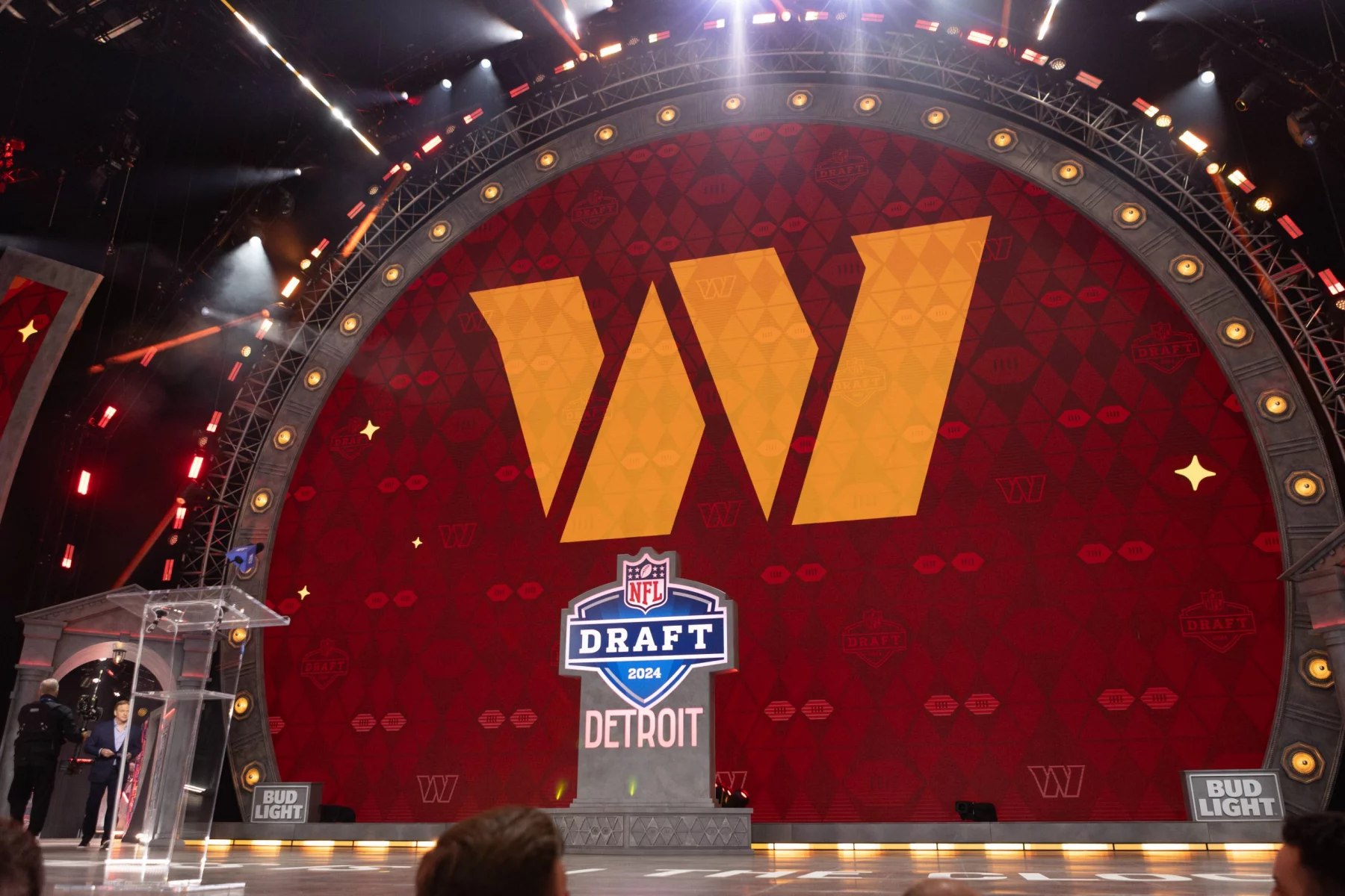 NFL Draft 2027 - Auf einer großen Bühne prangt ein riesiges gelbes „W“-Logo auf rotem Grund. Darunter sind das NFL Draft 2024-Schild und „DETROIT“ zu sehen. Die Bühne ist mit hellen Scheinwerfern beleuchtet. Links neben einem durchsichtigen Podium stehen Menschen; in den Ecken sind Bud-Light-Logos zu sehen. Diese Beschreibung wurde mit der FootballR KI automatisch generiert.