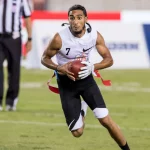 Darrell Doucette - Ein Flag-Football-Spieler in weißem ärmellosem Trikot und schwarzen Shorts läuft mit einem Football in der Hand über einen Rasenplatz. Er trägt eine rote Flagge an der Hüfte, seine langen Haare sind zurückgebunden und er wirkt konzentriert. Im Hintergrund sind ein Schiedsrichter und Werbespots zu sehen. Diese Beschreibung wurde mit der FootballR KI automatisch generiert.