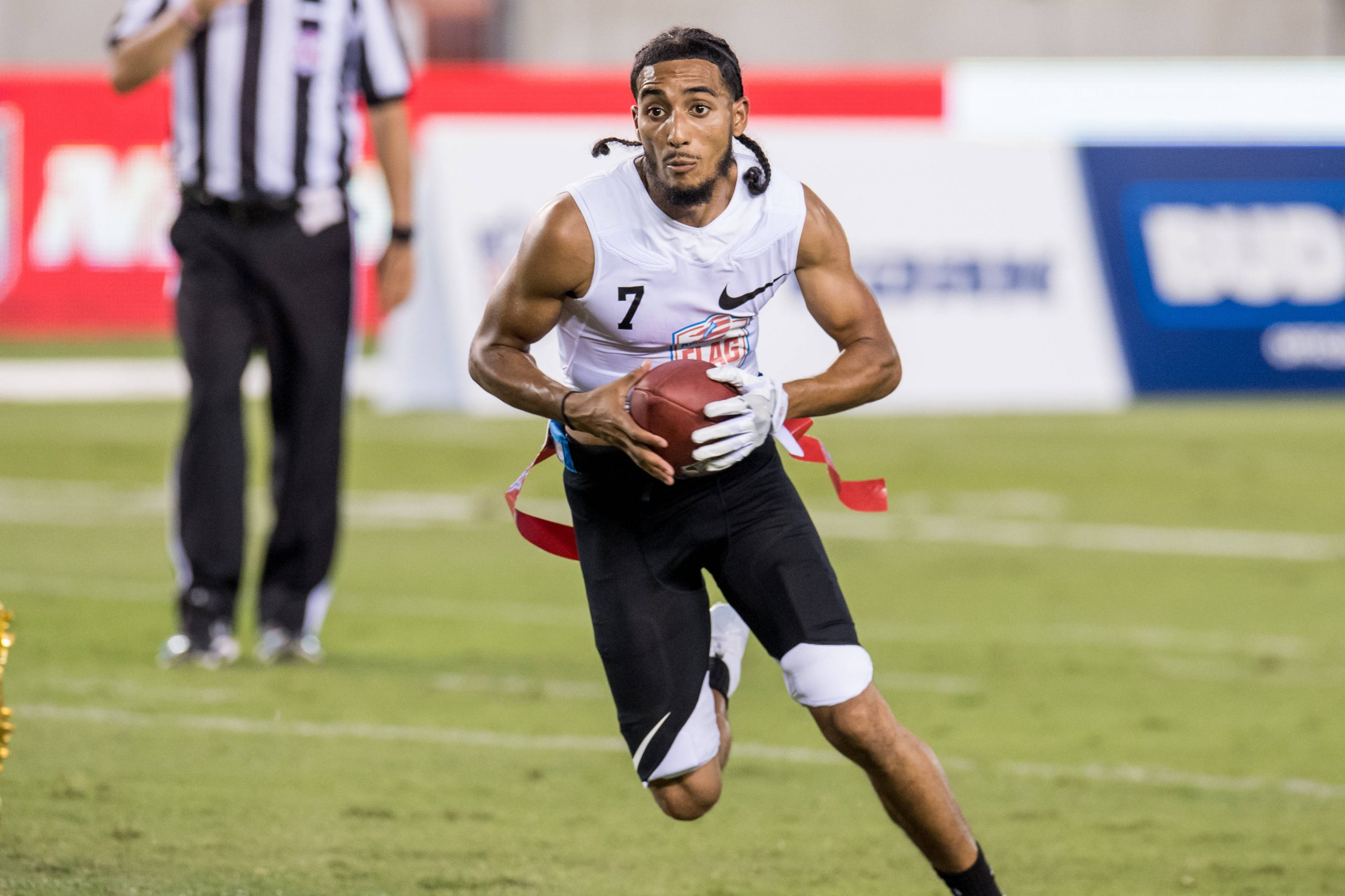 Darrell Doucette - Ein Flag-Football-Spieler in weißem ärmellosem Trikot und schwarzen Shorts läuft mit einem Football in der Hand über einen Rasenplatz. Er trägt eine rote Flagge an der Hüfte, seine langen Haare sind zurückgebunden und er wirkt konzentriert. Im Hintergrund sind ein Schiedsrichter und Werbespots zu sehen. Diese Beschreibung wurde mit der FootballR KI automatisch generiert.