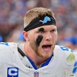 Aidan Hutchinson, ein Footballspieler der Detroit Lions mit schwarzem Stirnband, schwarzer Augenfarbe und schmutzig-weißer Uniform, schreit während eines Spiels lautstark. Der Stadionhintergrund ist unscharf, was seinen emotionalen und kämpferischen Ausdruck unterstreicht. Diese Beschreibung wurde mit der FootballR KI automatisch generiert.