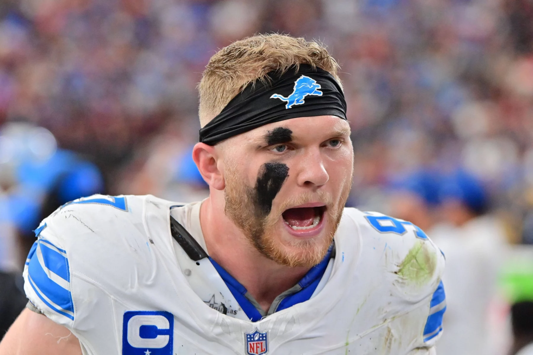Aidan Hutchinson, ein Footballspieler der Detroit Lions mit schwarzem Stirnband, schwarzer Augenfarbe und schmutzig-weißer Uniform, schreit während eines Spiels lautstark. Der Stadionhintergrund ist unscharf, was seinen emotionalen und kämpferischen Ausdruck unterstreicht. Diese Beschreibung wurde mit der FootballR KI automatisch generiert.