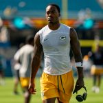 Ein muskulöser Packers Footballspieler in einem ärmellosen Green Bay Packers-Shirt und gelben Shorts läuft mit Kopfhörern über das Spielfeld. Mit Tattoos an beiden Armen und einem konzentrierten Gesichtsausdruck fällt er vor den verschwommenen Stadionsitzen auf. Diese Beschreibung wurde mit der FootballR KI automatisch generiert.