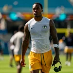 Ein muskulöser Packers Footballspieler in einem ärmellosen Green Bay Packers-Shirt und gelben Shorts läuft mit Kopfhörern über das Spielfeld. Mit Tattoos an beiden Armen und einem konzentrierten Gesichtsausdruck fällt er vor den verschwommenen Stadionsitzen auf. Diese Beschreibung wurde mit der FootballR KI automatisch generiert.