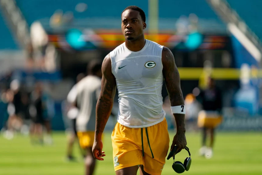 Ein muskulöser Packers Footballspieler in einem ärmellosen Green Bay Packers-Shirt und gelben Shorts läuft mit Kopfhörern über das Spielfeld. Mit Tattoos an beiden Armen und einem konzentrierten Gesichtsausdruck fällt er vor den verschwommenen Stadionsitzen auf. Diese Beschreibung wurde mit der FootballR KI automatisch generiert.