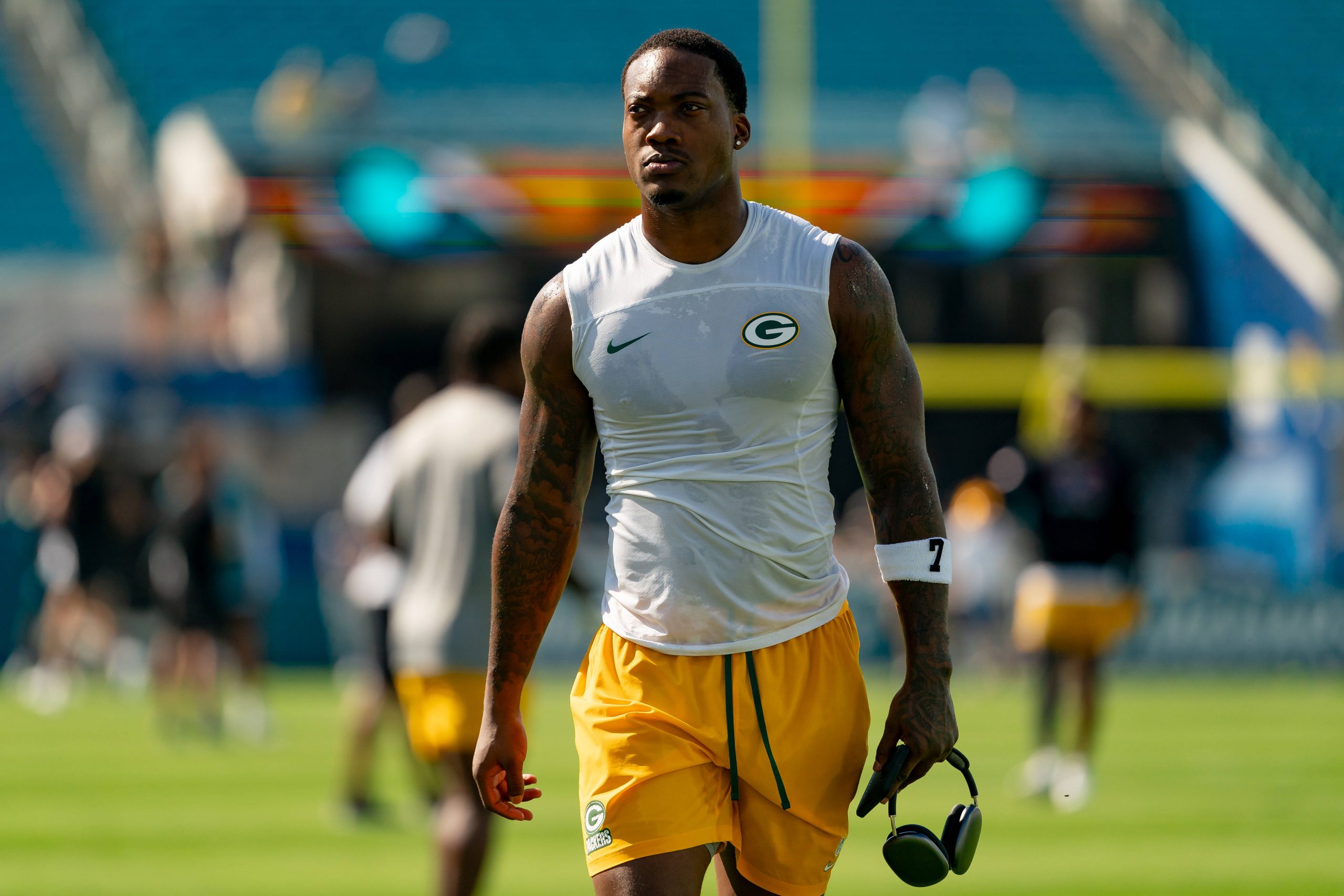 Ein muskulöser Packers Footballspieler in einem ärmellosen Green Bay Packers-Shirt und gelben Shorts läuft mit Kopfhörern über das Spielfeld. Mit Tattoos an beiden Armen und einem konzentrierten Gesichtsausdruck fällt er vor den verschwommenen Stadionsitzen auf. Diese Beschreibung wurde mit der FootballR KI automatisch generiert.