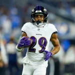 Ein Footballspieler der Baltimore Ravens mit der Nummer 29, in weißem Trikot, violetten Handschuhen und Helm, läuft mit dem Ball über das Feld – bemerkenswerte Widerstandskraft nach der Genesung von einem Achillessehnenriss. Der Hintergrund ist unscharf und zeigt andere Spieler und Zuschauer. Diese Beschreibung wurde mit der FootballR KI automatisch generiert.