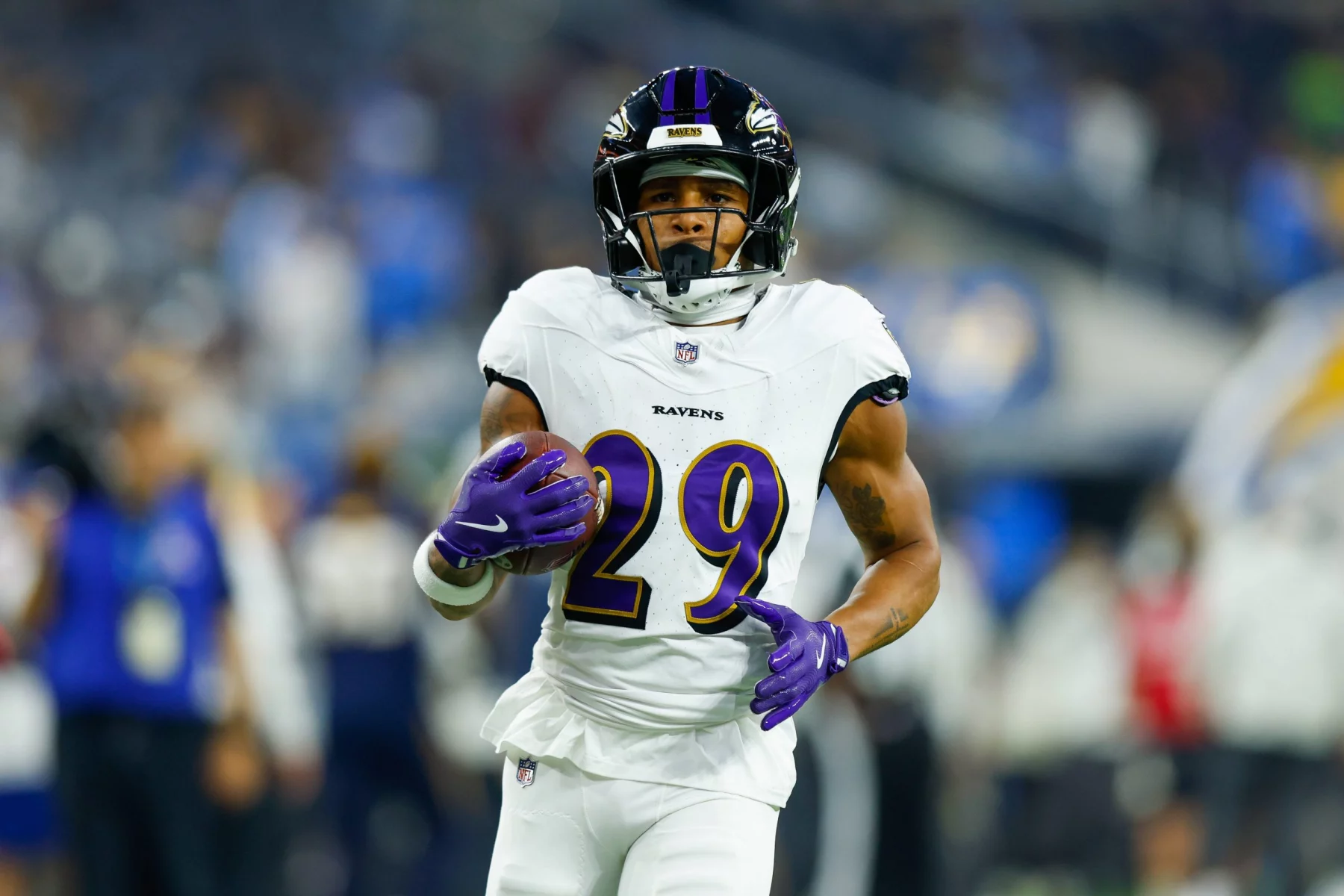 Ein Footballspieler der Baltimore Ravens mit der Nummer 29, in weißem Trikot, violetten Handschuhen und Helm, läuft mit dem Ball über das Feld – bemerkenswerte Widerstandskraft nach der Genesung von einem Achillessehnenriss. Der Hintergrund ist unscharf und zeigt andere Spieler und Zuschauer. Diese Beschreibung wurde mit der FootballR KI automatisch generiert.