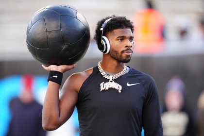 Nummer 12 - Shedeur Sanders trägt ein schwarzes langärmeliges Nike-Shirt und große Kopfhörer und hält einen schwarzen Medizinball auf der Schulter. Er hat einen gestutzten Bart, kurzes lockiges Haar und eine dicke, juwelenbesetzte Kette. Er steht vor verschwommenen Stadionsitzen und unscharfen Figuren. Diese Beschreibung wurde mit der FootballR KI automatisch generiert.
