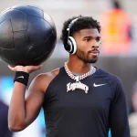 Nummer 12 - Shedeur Sanders trägt ein schwarzes langärmeliges Nike-Shirt und große Kopfhörer und hält einen schwarzen Medizinball auf der Schulter. Er hat einen gestutzten Bart, kurzes lockiges Haar und eine dicke, juwelenbesetzte Kette. Er steht vor verschwommenen Stadionsitzen und unscharfen Figuren. Diese Beschreibung wurde mit der FootballR KI automatisch generiert.