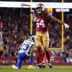 Fred Warner, ein Spieler der San Francisco 49ers im roten Trikot (#54), spannt beim Jubeln auf dem Spielfeld seinen Arm an. Links steht ein Teamkollege, daneben kniet ein Spieler der Los Angeles Rams (#23). Die Menge füllt das Stadion unter hellem Licht. Diese Beschreibung wurde mit der FootballR KI automatisch generiert.