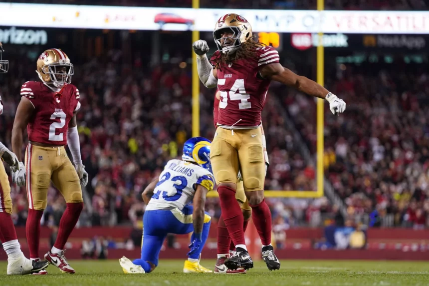 Fred Warner, ein Spieler der San Francisco 49ers im roten Trikot (#54), spannt beim Jubeln auf dem Spielfeld seinen Arm an. Links steht ein Teamkollege, daneben kniet ein Spieler der Los Angeles Rams (#23). Die Menge füllt das Stadion unter hellem Licht. Diese Beschreibung wurde mit der FootballR KI automatisch generiert.