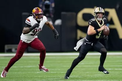 Jake Haener, Quarterback der New Orleans Saints, hält den Ball und will passen. Während eines spannenden Spiels rennt er nach vorn, während ihn ein Verteidiger der Washington Commanders auf dem grünen Footballfeld verfolgt. Beide Spieler sind in voller Uniform und es herrscht intensive Action. Diese Beschreibung wurde mit der FootballR KI automatisch generiert.