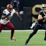 Jake Haener, Quarterback der New Orleans Saints, hält den Ball und will passen. Während eines spannenden Spiels rennt er nach vorn, während ihn ein Verteidiger der Washington Commanders auf dem grünen Footballfeld verfolgt. Beide Spieler sind in voller Uniform und es herrscht intensive Action. Diese Beschreibung wurde mit der FootballR KI automatisch generiert.