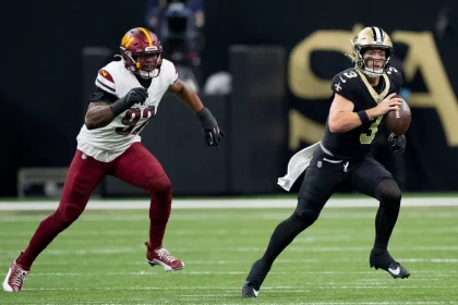 Jake Haener, Quarterback der New Orleans Saints, hält den Ball und will passen. Während eines spannenden Spiels rennt er nach vorn, während ihn ein Verteidiger der Washington Commanders auf dem grünen Footballfeld verfolgt. Beide Spieler sind in voller Uniform und es herrscht intensive Action. Diese Beschreibung wurde mit der FootballR KI automatisch generiert.