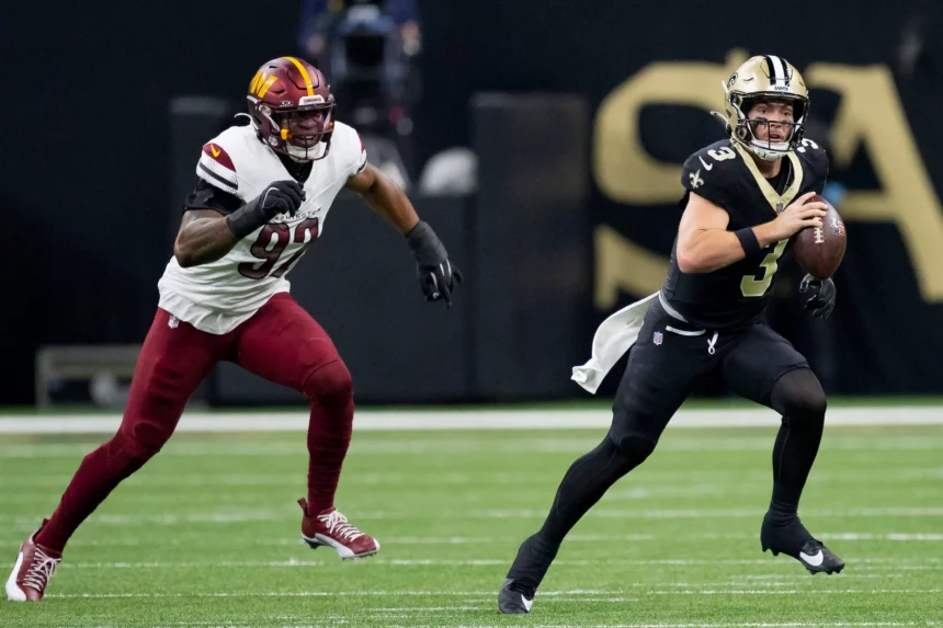 Jake Haener, Quarterback der New Orleans Saints, hält den Ball und will passen. Während eines spannenden Spiels rennt er nach vorn, während ihn ein Verteidiger der Washington Commanders auf dem grünen Footballfeld verfolgt. Beide Spieler sind in voller Uniform und es herrscht intensive Action. Diese Beschreibung wurde mit der FootballR KI automatisch generiert.