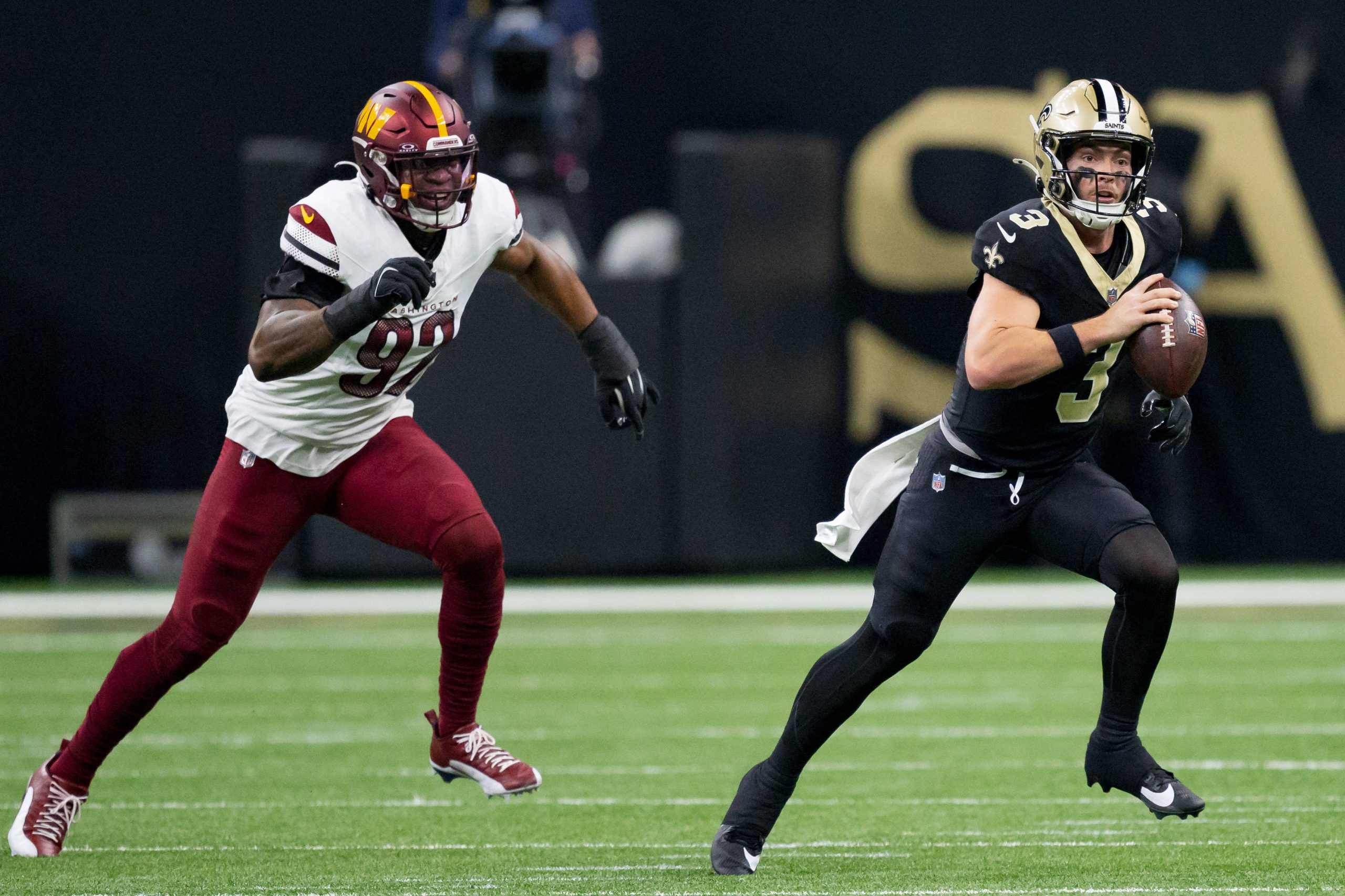 Jake Haener, Quarterback der New Orleans Saints, hält den Ball und will passen. Während eines spannenden Spiels rennt er nach vorn, während ihn ein Verteidiger der Washington Commanders auf dem grünen Footballfeld verfolgt. Beide Spieler sind in voller Uniform und es herrscht intensive Action. Diese Beschreibung wurde mit der FootballR KI automatisch generiert.