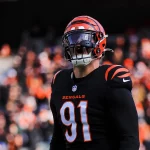 Trey Hendrickson, der die Nummer 91 in Trikot und Helm der Cincinnati Bengals trägt, steht konzentriert auf dem Spielfeld. Die schwarz-orangefarbene Ausrüstung mit sichtbaren Bengals-Logos bildet einen Kontrast zum verschwommenen Bild des mit Fans gefüllten Stadions. Diese Beschreibung wurde mit der FootballR KI automatisch generiert.