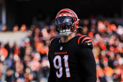 Trey Hendrickson, der die Nummer 91 in Trikot und Helm der Cincinnati Bengals trägt, steht konzentriert auf dem Spielfeld. Die schwarz-orangefarbene Ausrüstung mit sichtbaren Bengals-Logos bildet einen Kontrast zum verschwommenen Bild des mit Fans gefüllten Stadions. Diese Beschreibung wurde mit der FootballR KI automatisch generiert.
