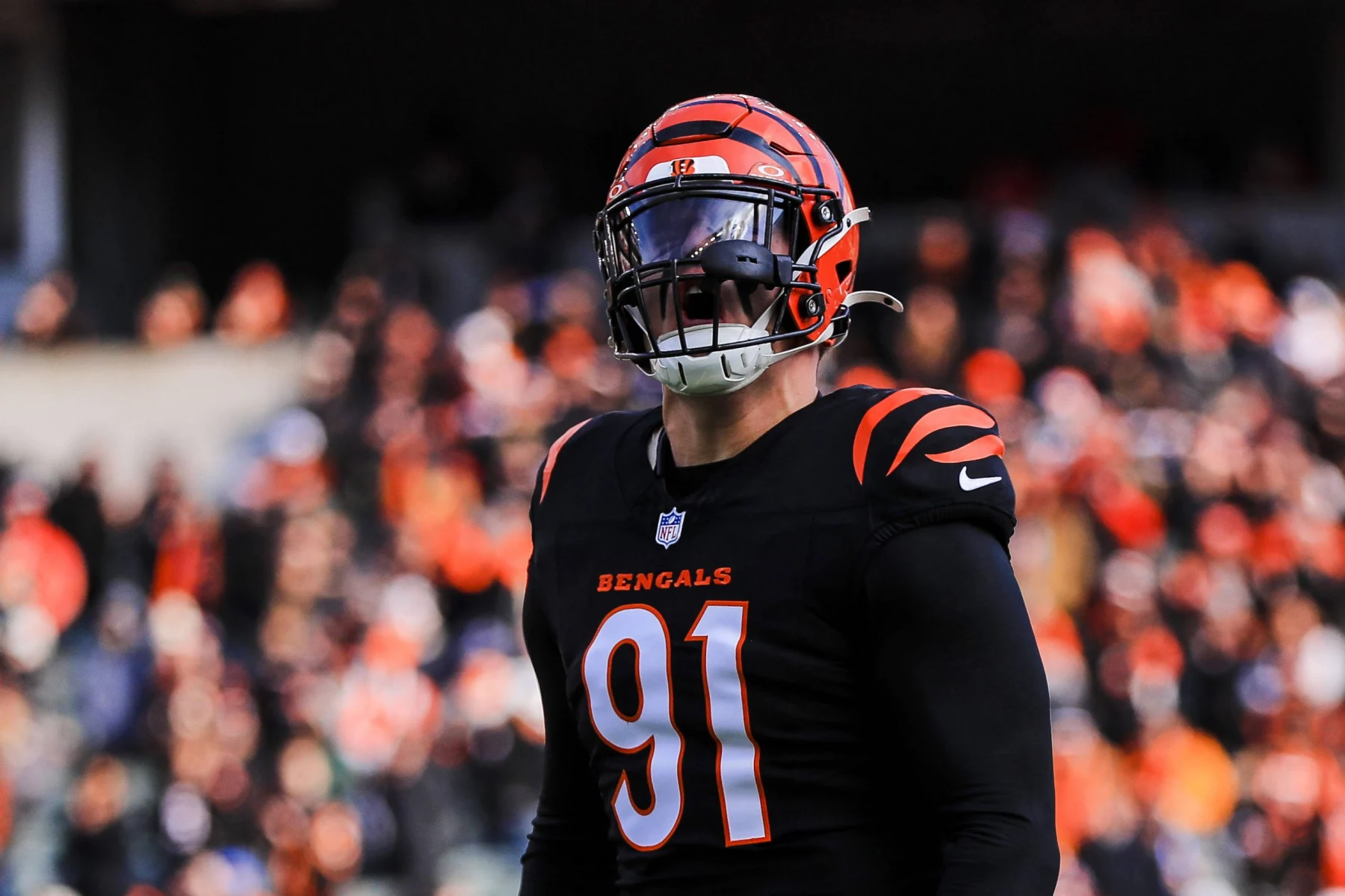 Trey Hendrickson, der die Nummer 91 in Trikot und Helm der Cincinnati Bengals trägt, steht konzentriert auf dem Spielfeld. Die schwarz-orangefarbene Ausrüstung mit sichtbaren Bengals-Logos bildet einen Kontrast zum verschwommenen Bild des mit Fans gefüllten Stadions. Diese Beschreibung wurde mit der FootballR KI automatisch generiert.