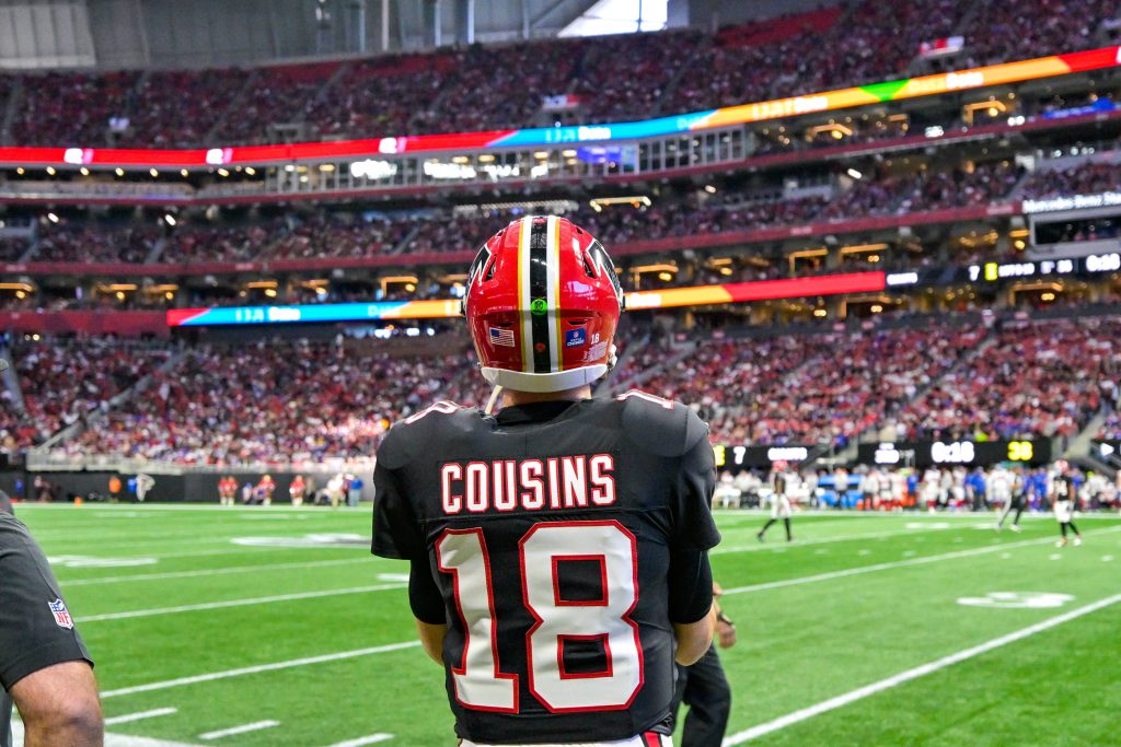 Kirk Cousins - Ein American-Football-Spieler in einem schwarzen Trikot mit der Aufschrift „COUSINS“ und der Nummer 18 steht auf dem Spielfeld und blickt auf ein überfülltes Stadion – vielleicht denkt er gerade über seinen nächsten großen Cousins-Deal nach. Fans füllen die Tribüne, im Hintergrund sind weitere Spieler zu sehen. Diese Beschreibung wurde mit der FootballR KI automatisch generiert.