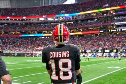 Kirk Cousins - Ein American-Football-Spieler in einem schwarzen Trikot mit der Aufschrift „COUSINS“ und der Nummer 18 steht auf dem Spielfeld und blickt auf ein überfülltes Stadion – vielleicht denkt er gerade über seinen nächsten großen Cousins-Deal nach. Fans füllen die Tribüne, im Hintergrund sind weitere Spieler zu sehen. Diese Beschreibung wurde mit der FootballR KI automatisch generiert.