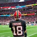 Kirk Cousins - Ein American-Football-Spieler in einem schwarzen Trikot mit der Aufschrift „COUSINS“ und der Nummer 18 steht auf dem Spielfeld und blickt auf ein überfülltes Stadion – vielleicht denkt er gerade über seinen nächsten großen Cousins-Deal nach. Fans füllen die Tribüne, im Hintergrund sind weitere Spieler zu sehen. Diese Beschreibung wurde mit der FootballR KI automatisch generiert.