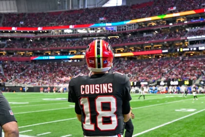 Kirk Cousins - Ein American-Football-Spieler in einem schwarzen Trikot mit der Aufschrift „COUSINS“ und der Nummer 18 steht auf dem Spielfeld und blickt auf ein überfülltes Stadion – vielleicht denkt er gerade über seinen nächsten großen Cousins-Deal nach. Fans füllen die Tribüne, im Hintergrund sind weitere Spieler zu sehen. Diese Beschreibung wurde mit der FootballR KI automatisch generiert.