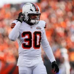 Devin Bush, der eine weiße Uniform mit orange-braunen Streifen und der Nummer 30 trägt, steht auf dem Spielfeld und rückt seinen Helm zurecht. Die verschwommene Menge im Hintergrund ist überwiegend in Orange und Schwarz gekleidet. Diese Beschreibung wurde mit der FootballR KI automatisch generiert.