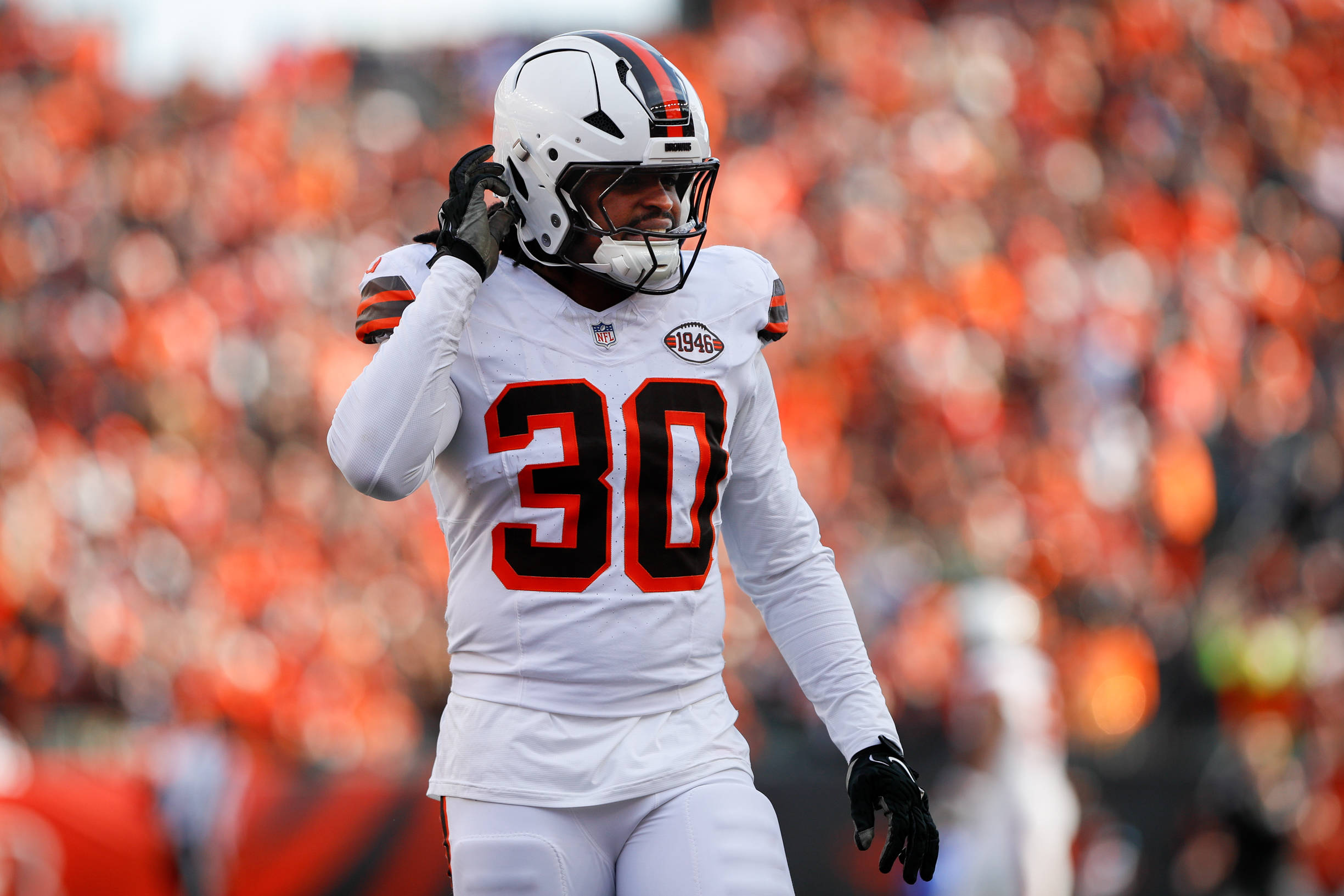 Devin Bush, der eine weiße Uniform mit orange-braunen Streifen und der Nummer 30 trägt, steht auf dem Spielfeld und rückt seinen Helm zurecht. Die verschwommene Menge im Hintergrund ist überwiegend in Orange und Schwarz gekleidet. Diese Beschreibung wurde mit der FootballR KI automatisch generiert.