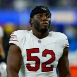 Victor Dimukeje steht im weißen Arizona-Cardinals-Trikot mit der Nummer 52 auf dem Spielfeld. Er trägt ein schwarzes Stirnband, Schulterpolster und schwarze Gesichtsbemalung unter den Augen. Im unscharfen Hintergrund sind ein weiterer Spieler und blaue Stadionlichter zu sehen. Diese Beschreibung wurde mit der FootballR KI automatisch generiert.