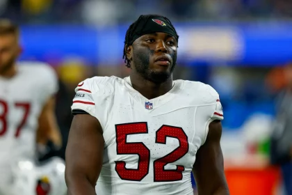 Victor Dimukeje steht im weißen Arizona-Cardinals-Trikot mit der Nummer 52 auf dem Spielfeld. Er trägt ein schwarzes Stirnband, Schulterpolster und schwarze Gesichtsbemalung unter den Augen. Im unscharfen Hintergrund sind ein weiterer Spieler und blaue Stadionlichter zu sehen. Diese Beschreibung wurde mit der FootballR KI automatisch generiert.