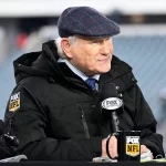 Terry Bradshaw mit blaukarierter Schiebermütze und schwarzer Jacke mit dem „FOX NFL“-Logo sitzt mit einem Mikrofon und einer Tasse von FOX Sports an einem Schreibtisch und lächelt leicht. Hinter ihm verschwimmen die leeren Stadionsitze, während Terry Bradshaw die Quarterback-Strategie der Steelers kritisiert. Diese Beschreibung wurde mit der FootballR KI automatisch generiert.