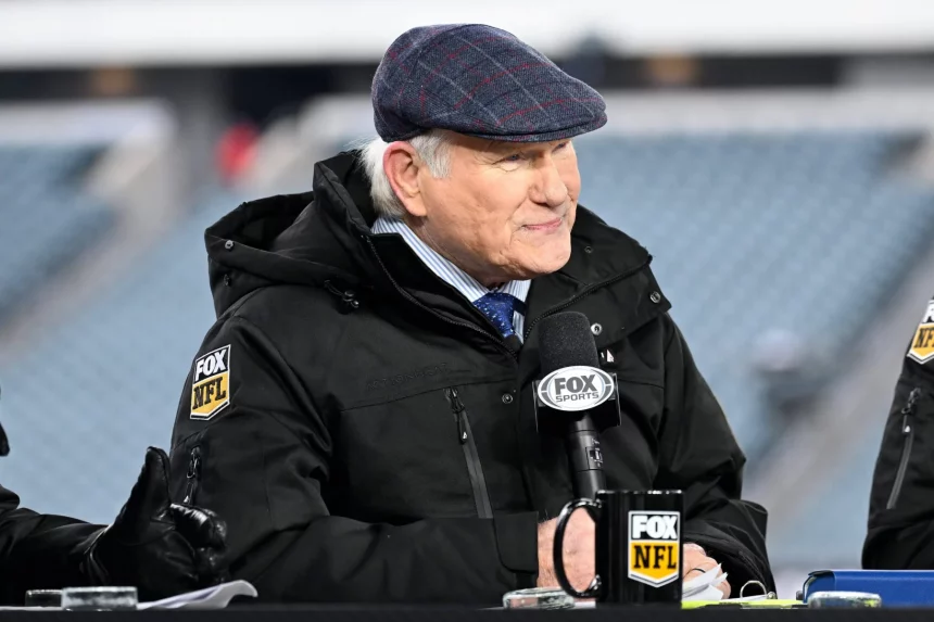 Terry Bradshaw mit blaukarierter Schiebermütze und schwarzer Jacke mit dem „FOX NFL“-Logo sitzt mit einem Mikrofon und einer Tasse von FOX Sports an einem Schreibtisch und lächelt leicht. Hinter ihm verschwimmen die leeren Stadionsitze, während Terry Bradshaw die Quarterback-Strategie der Steelers kritisiert. Diese Beschreibung wurde mit der FootballR KI automatisch generiert.