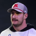 Ein Mann mit hellem Bart, der Joey Bosa ähnelt, trägt eine grau-weiße Kappe mit einem roten „A“-Logo und ein weißes Hemd über einem schwarzen Kapuzenpullover. Er blickt leicht zur Seite und steht vor einem dunklen, unscharfen Hintergrund mit sanfter Beleuchtung. Diese Beschreibung wurde mit der FootballR KI automatisch generiert.