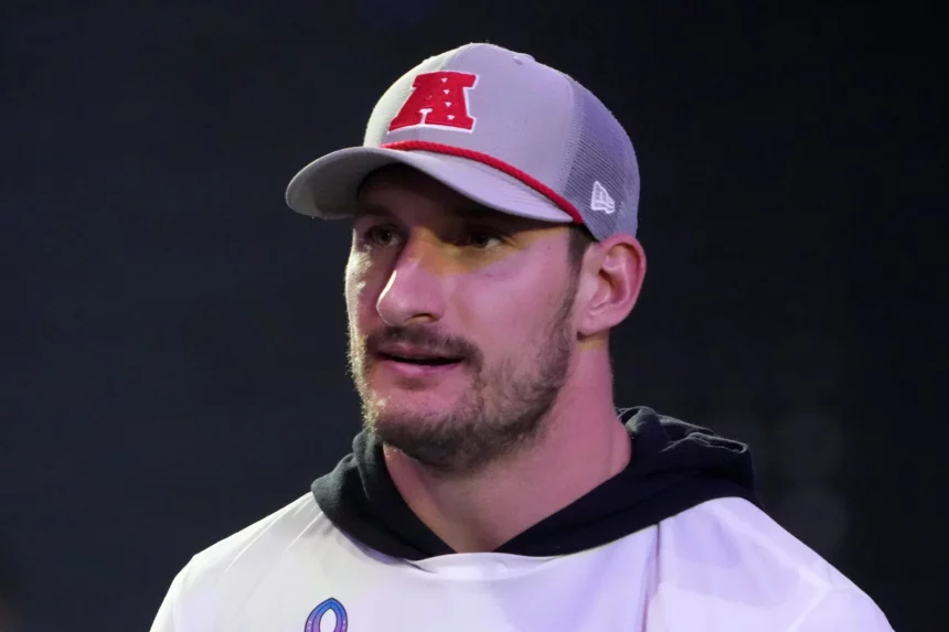 Ein Mann mit hellem Bart, der Joey Bosa ähnelt, trägt eine grau-weiße Kappe mit einem roten „A“-Logo und ein weißes Hemd über einem schwarzen Kapuzenpullover. Er blickt leicht zur Seite und steht vor einem dunklen, unscharfen Hintergrund mit sanfter Beleuchtung. Diese Beschreibung wurde mit der FootballR KI automatisch generiert.