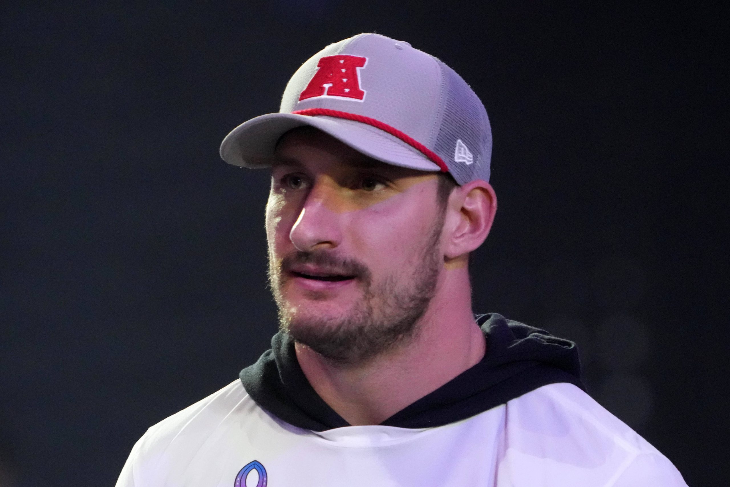 Ein Mann mit hellem Bart, der Joey Bosa ähnelt, trägt eine grau-weiße Kappe mit einem roten „A“-Logo und ein weißes Hemd über einem schwarzen Kapuzenpullover. Er blickt leicht zur Seite und steht vor einem dunklen, unscharfen Hintergrund mit sanfter Beleuchtung. Diese Beschreibung wurde mit der FootballR KI automatisch generiert.