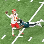 Heute im TV - Route Concepts - Ein Footballspieler in der roten Uniform der Kansas City Chiefs (#15) bereitet sich darauf vor, den Ball zu werfen und demonstriert dabei scharfe Routenkonzepte, während er von einem grünen Spieler der Philadelphia Eagles (#94) auf einem grünen Feld mit weißen Yard-Linien getackelt wird. Diese Beschreibung wurde mit der FootballR KI automatisch generiert.