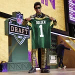 Ein lächelnder Matthew Golden in einem goldgemusterten Anzug und Sonnenbrille hält auf der Bühne beim NFL Draft 2025 ein grünes Packers-Trikot mit der Aufschrift „GOLDEN 1“ hoch. Das NFL-Draft-Logo und das „Bud Light“-Schild sind zu sehen, eine weitere Person geht vorbei. Diese Beschreibung wurde mit der FootballR KI automatisch generiert.