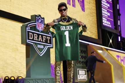 Ein lächelnder Matthew Golden in einem goldgemusterten Anzug und Sonnenbrille hält auf der Bühne beim NFL Draft 2025 ein grünes Packers-Trikot mit der Aufschrift „GOLDEN 1“ hoch. Das NFL-Draft-Logo und das „Bud Light“-Schild sind zu sehen, eine weitere Person geht vorbei. Diese Beschreibung wurde mit der FootballR KI automatisch generiert.