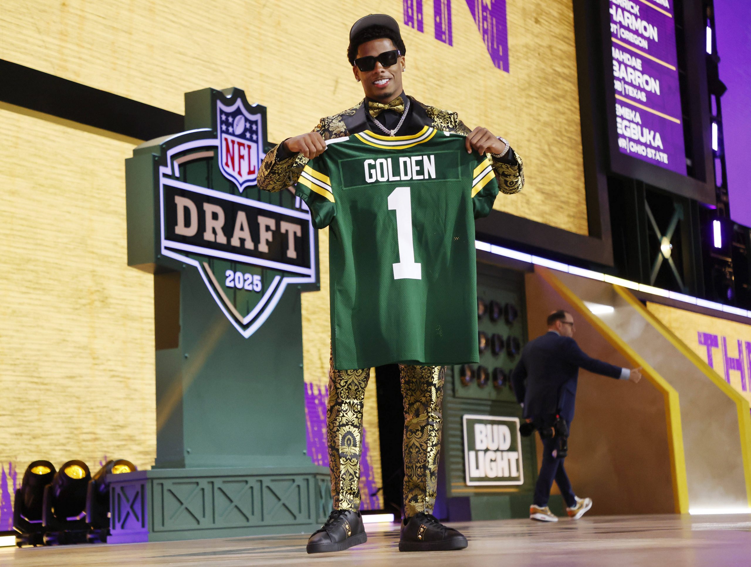 Ein lächelnder Matthew Golden in einem goldgemusterten Anzug und Sonnenbrille hält auf der Bühne beim NFL Draft 2025 ein grünes Packers-Trikot mit der Aufschrift „GOLDEN 1“ hoch. Das NFL-Draft-Logo und das „Bud Light“-Schild sind zu sehen, eine weitere Person geht vorbei. Diese Beschreibung wurde mit der FootballR KI automatisch generiert.