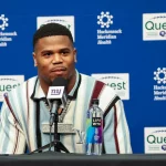 Abdul Carter sitzt an einem Tisch mit einem Mikrofon der New York Giants. Er trägt ein gestreiftes Hemd, das dezent auf seine Trikotnummer hindeutet, und eine große Halskette. Vor ihm steht eine Flasche LIFEWTR-Wasser. Im Hintergrund sind die Logos von Hackensack Meridian Health und Quest zu sehen. Diese Beschreibung wurde mit der FootballR KI automatisch generiert.