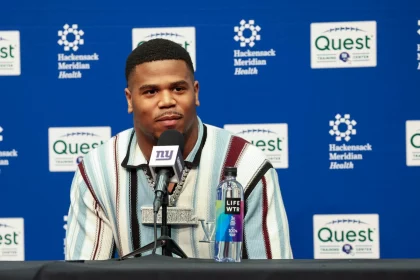 Abdul Carter sitzt an einem Tisch mit einem Mikrofon der New York Giants. Er trägt ein gestreiftes Hemd, das dezent auf seine Trikotnummer hindeutet, und eine große Halskette. Vor ihm steht eine Flasche LIFEWTR-Wasser. Im Hintergrund sind die Logos von Hackensack Meridian Health und Quest zu sehen. Diese Beschreibung wurde mit der FootballR KI automatisch generiert.