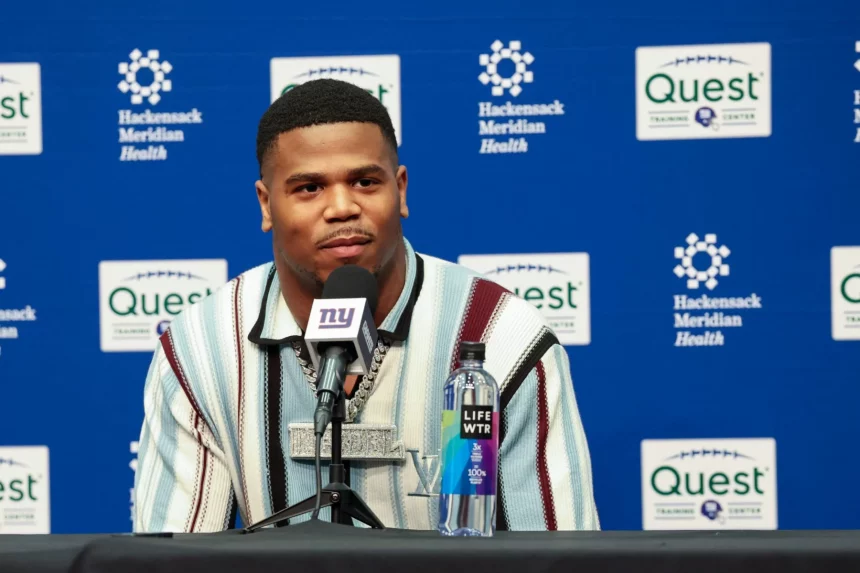 Abdul Carter sitzt an einem Tisch mit einem Mikrofon der New York Giants. Er trägt ein gestreiftes Hemd, das dezent auf seine Trikotnummer hindeutet, und eine große Halskette. Vor ihm steht eine Flasche LIFEWTR-Wasser. Im Hintergrund sind die Logos von Hackensack Meridian Health und Quest zu sehen. Diese Beschreibung wurde mit der FootballR KI automatisch generiert.